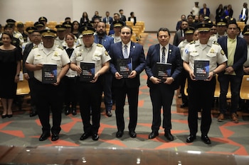 Cinco hombres, tres con uniforme policial y dos con traje, sostienen libros en un auditorio con más personas y sillas al fondo