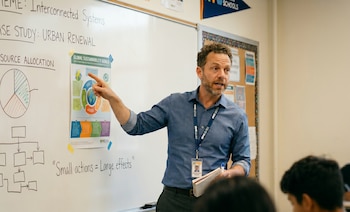 Un profesor barbudo con camisa azul y credencial de NYC DOE señala un póster de Objetivos de Sostenibilidad Globales en una pizarra blanca de aula