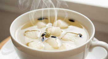 Primer plano de una taza de té blanco humeante con cubitos de pera y semillas de cardamomo flotando en el líquido, bajo una suave luz natural desde la derecha.