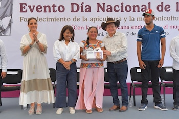 Grupo de personas en un escenario con un telón de fondo. Una mujer recibe un paquete de un hombre mientras otra mujer aplaude. Otros hombres de pie
