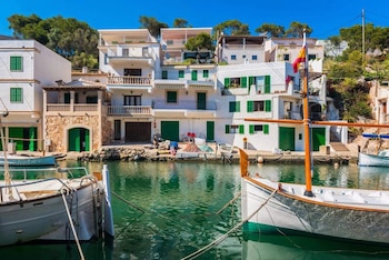Puerto natural con casas blancas, persianas verdes y balcones. Varias embarcaciones atracadas en el agua turquesa. Vegetación y cielo azul