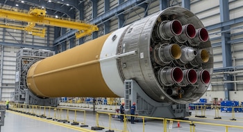 Vista de la etapa central horizontal del cohete Space Launch System (SLS) dentro de un gran hangar, con múltiples motores visibles en un extremo y dos técnicos trabajando cerca.