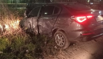 Un automóvil de color gris oscuro con daños en su lateral reposa sobre la vegetación a un costado del camino durante la noche