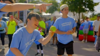 Un joven con camiseta azul y pelo oscuro sonríe mientras sostiene una pelota verde. Detrás hay otras personas y árboles en un día soleado