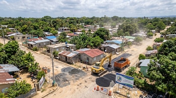 Vista aérea de calle en Los Alcarrizos siendo pavimentada. Se ve una excavadora, camión volquete, asfalto, casas, vegetación y un letrero municipal.