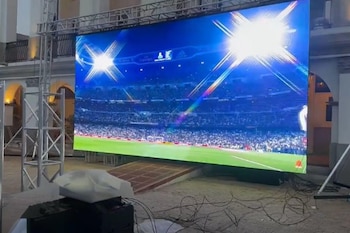 Una pantalla LED gigante al aire libre, montada sobre una estructura de metal, reproduce un partido de fútbol en un estadio iluminado