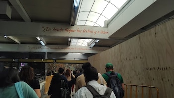 Varias personas caminan por un pasillo de la estación del Metro con barricadas de madera a la derecha y un techo translúcido con textos en rojo