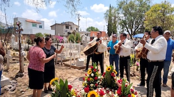 Un mariachi con instrumentos y varias personas se reúnen en un panteón mexicano alrededor de una tumba decorada con flores rojas y amarillas bajo cielo soleado.