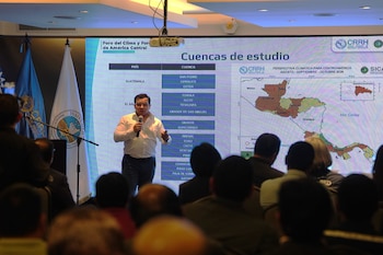 Hombre habla por micrófono ante pantalla gigante con mapa de Centroamérica y listado de cuencas, banderas institucionales y público