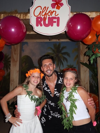 Un hombre sonríe abrazando a dos niñas vestidas con atuendos festivos; de fondo hay globos, un cartel y una escenografía tropical