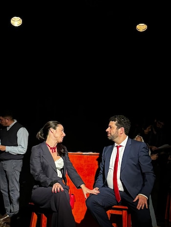 Dos actores, un hombre con traje azul y corbata roja, y una mujer con traje oscuro y collar rojo, sentados en sillas rojas, se miran en un escenario oscuro