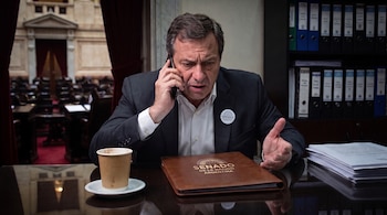 Un hombre con traje habla por celular ante una carpeta con la leyenda del Senado de la Nación Argentina, un vaso de café y pilas de hojas.