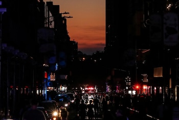 Calle de Nueva York en la oscuridad con siluetas de peatones, luces de vehículos y luces rojas intermitentes de emergencia. El cielo es naranja rojizo