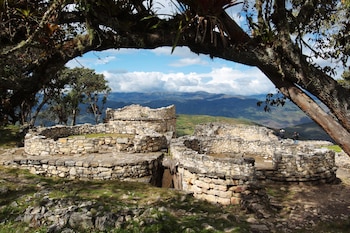 Ruinas arqueológicas de piedra circulares, árboles con musgo y bromelias, montañas en el horizonte, cielo con nubes, dos personas observan el paisaje