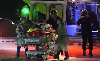 Cuatro personas con indumentaria de emergencia trasladan una camilla con un paciente hacia un vehículo de emergencia con luces verdes y rojas. Hay un símbolo A en el suelo