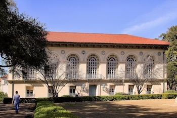 Un hombre camina por un sendero frente a Battle Hall, un edificio de piedra clara con techo de tejas rojas, grandes ventanas arqueadas y un jardín delantero