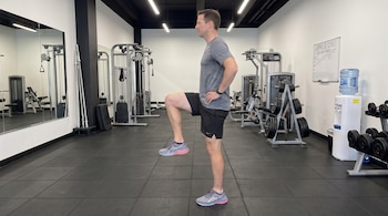 Hombre de perfil con ropa deportiva y una pierna flexionada a noventa grados dentro de un gimnasio con máquinas de pesas