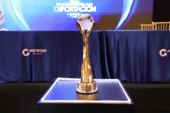 Trofeo dorado con un globo de cristal en la cima sobre un pedestal iluminado. Al fondo, una pantalla con texto "EXPORTACION 2020" y un logotipo AGEXPORT