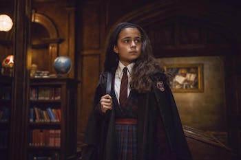 Una niña vestida con túnica oscura y corbata permanece de pie en una habitación con estantes con libros y un globo terráqueo