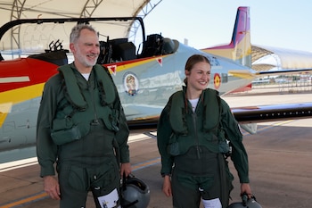 Hombre con barba y mujer joven, ambos con trajes de vuelo verdes y chalecos salvavidas, sostienen cascos y sonríen frente a un avión militar