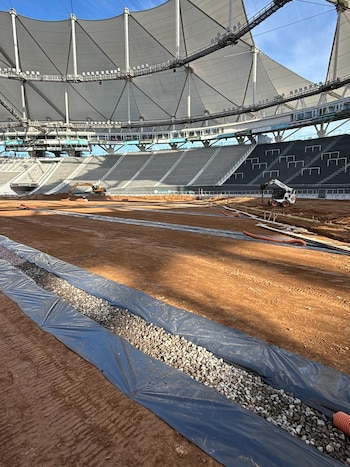 Vista interior de un estadio en remodelación con el campo de tierra, vehículos de construcción, gradas vacías y techo de tela tensada bajo cielo azul
