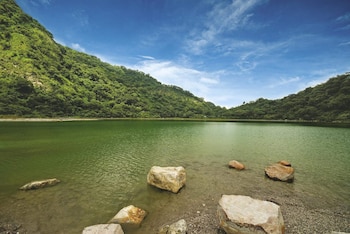 Vista panorámica de un lago de agua verde, con grandes rocas en la orilla, rodeado de montañas cubiertas de vegetación bajo un cielo azul nublado