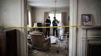 Escena de un crimen en una sala de estar donde policías y forenses examinan un cuerpo cubierto. Hay cinta policial y un retrato de una mujer mayor.