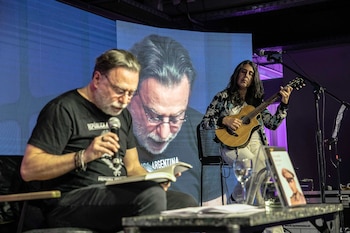 Un hombre lee un libro con un micrófono junto a una mujer que toca la guitarra frente a una pantalla en un escenario
