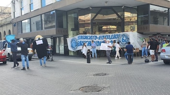 Calle urbana, edificio moderno, grupo de personas, banner Sindicato Hotelero Gastronómicos, letreros, megáfono, persona con disfraz, vehículos