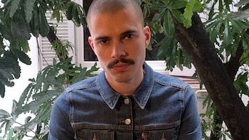 Hombre con cabello rapado, bigote y chaqueta vaquera azul mira hacia el frente, situado delante de plantas y una ventana