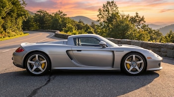 Vista lateral de un Porsche Carrera GT plateado estacionado en una carretera sinuosa. Al fondo, montañas verdes y un cielo anaranjado al atardecer.