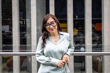 Mujer con cabello oscuro, gafas grandes y camisa verde claro. Se apoya en una barandilla de cristal. Detrás, fachada de edificio con ventanas y pilares