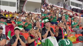 Una multitud de personas en las gradas de un estadio. Algunas visten camisetas verdes, otras sostienen banderas de México y varias aplauden.