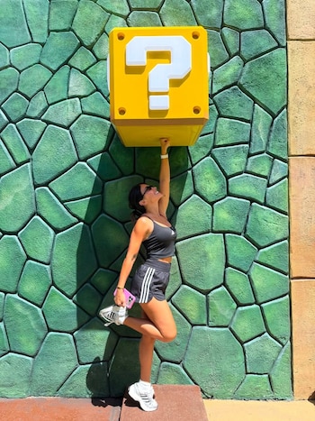 Mujer con top y shorts negros, zapatillas blancas, sostiene con una mano un gran bloque amarillo con signo de interrogación blanco, frente a una pared verde texturizada