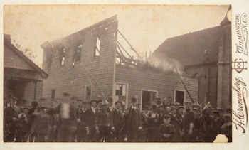Una fotografía antigua muestra un grupo de hombres posando frente a un edificio de madera en llamas o en ruinas, con humo visible. Otro edificio y texto aparecen en el borde derecho