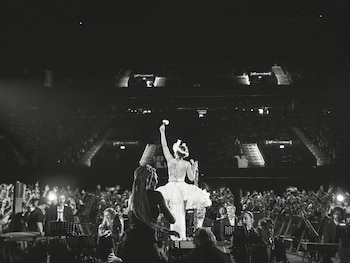 Cantante con vestido blanco y tocado levanta una flor en un escenario con músicos. Un público numeroso llena un estadio