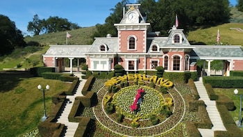 Vista aérea de la entrada principal de Neverland Ranch, un edificio de ladrillo rojo con una torre de reloj y banderas de EE.UU., rodeado de setos verdes y un gran reloj floral