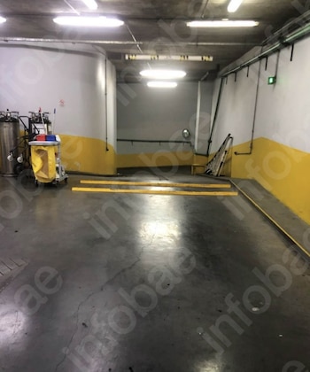 Imagen de un estacionamiento o área de servicio subterránea. El piso es de concreto, paredes blancas con franjas amarillas. Hay un carrito de limpieza y luces fluorescentes en el techo