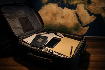 Una maleta abierta con un pasaporte de Estados Unidos, un teléfono móvil, documentos y un bolígrafo sobre una mesa junto a un mapa.
