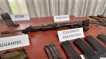 Mesa exhibe un fusil Galil, varias cacerinas abastecidas, una radio ICOM, guantes y carteles blancos de identificación de los objetos