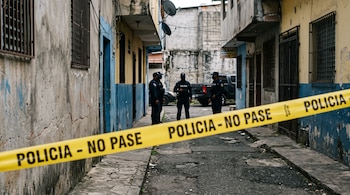 Una cinta amarilla con el texto POLICIA NO PASE atraviesa un callejón donde tres agentes policiales uniformados permanecen al fondo.
