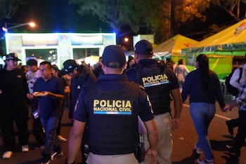 Dos agentes de policía con chalecos de "POLICIA NACIONAL CIVIL" caminan en medio de una multitud de personas, un arco iluminado y tiendas amarillas