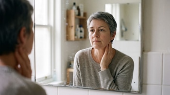Reflejo en espejo de mujer adulta con cabello gris corto, suéter claro y una mano en el cuello, frente a fondo desenfocado con ventana y estantería
