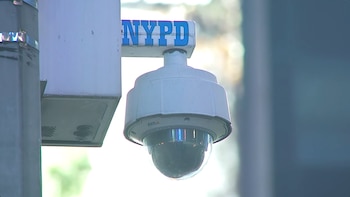 Primer plano de una cámara de seguridad domo de color blanco, con la marca 'Axis' visible, montada debajo de un letrero azul que dice 'NYPD' en un poste