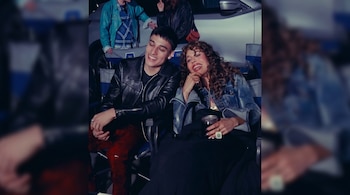 Hombre sonriente con chaqueta de cuero y pantalón rojo junto a mujer de pelo rizado y chaqueta de mezclilla sonriente, sentados en un lugar oscuro.