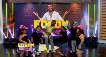 Edson Dávila sonríe en un escenario vibrante con luces de neón y confeti. Lo rodean cinco personas de baja estatura. Al fondo, una imagen grande de Dávila