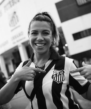 Fotografía en blanco y negro de una mujer sonriente que apunta con sus dedos hacia el escudo de su camiseta a rayas frente a un estadio
