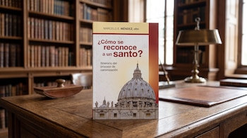 Un libro de pie sobre un escritorio de madera en una biblioteca con estanterías repletas de libros, una lámpara de mesa y un secante.