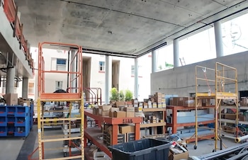 Interior de un edificio en construcción con techo de concreto sin terminar, andamios, cajas de materiales y gran entrada de luz natural a través de aberturas amplias