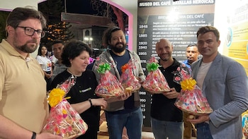 Grupo de cinco personas, tres hombres sonrientes y dos mujeres, sostienen canastas de regalo envueltas, con un letrero de 'MUSEO DEL CAFÉ' de fondo